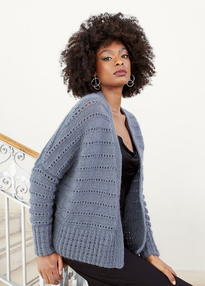 A person with curly hair wears a textured blue cardigan over a black outfit, posing confidently on a staircase with ornate railing. Neutral expression.