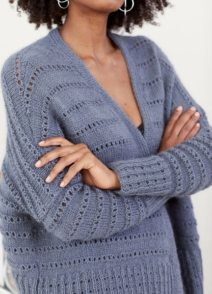 A person with curly hair and silver hoop earrings wears a textured, blue knit sweater. They stand with arms crossed, conveying confidence and style.