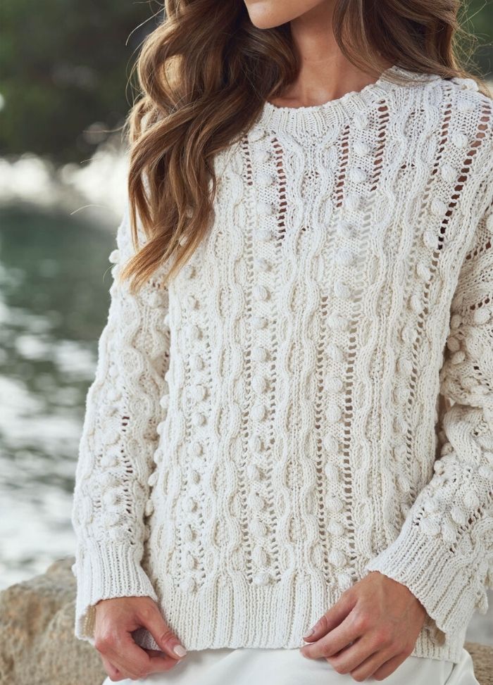 A woman wears a textured, cream cable-knit sweater, standing near a waterfront. Her wavy hair, relaxed stance, and soft light create a cozy, serene mood.