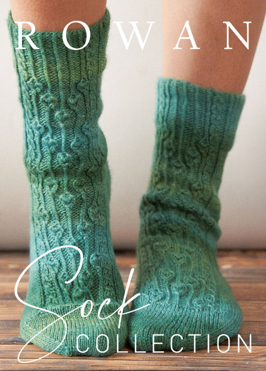 Two legs wearing textured, green knitted socks stand on a wooden floor. "Rowan Sock Collection" text overlays the image, suggesting a cozy, artisanal feel.