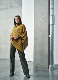 A woman stands confidently in a modern, industrial setting, wearing a mustard poncho and dark green pants. The concrete and metal background enhances the stylish, sophisticated mood.