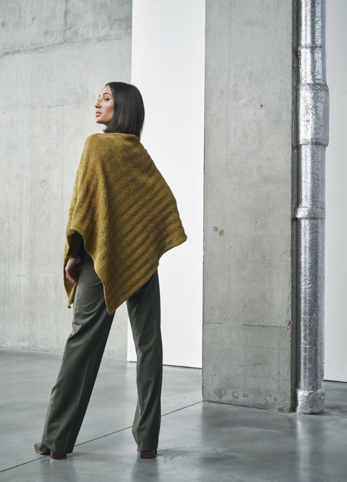 A woman stands in an industrial space wearing a mustard poncho and olive pants. She looks confident, with a neutral gray concrete wall backdrop.