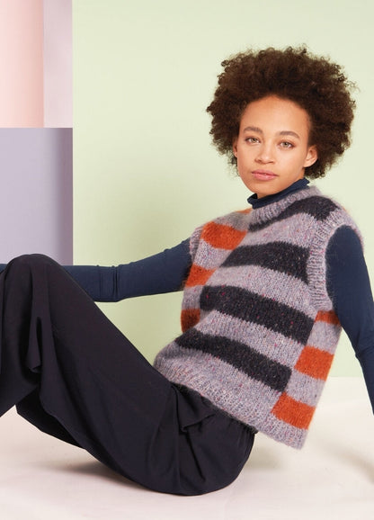 A person with curly hair leans back against a pastel geometric background. They wear a striped sleeveless sweater and dark pants, exuding a relaxed vibe.