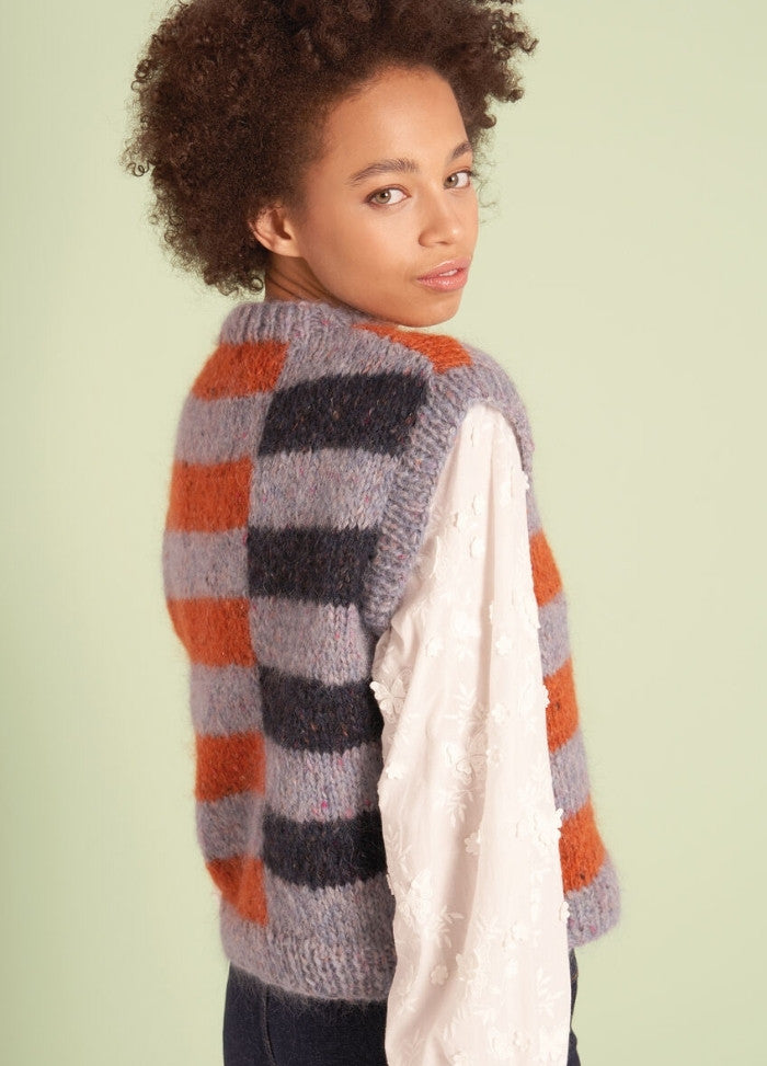 A young person with curly hair wearing a striped knit vest in orange, gray, and black over a white embroidered blouse, glancing over their shoulder.