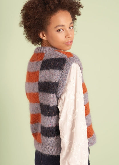 A young person with curly hair wearing a striped knit vest in orange, gray, and black over a white embroidered blouse, glancing over their shoulder.