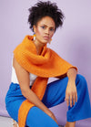 A woman with curly hair poses confidently in front of a lavender background. She wears a textured orange scarf, white top, and vibrant blue pants.