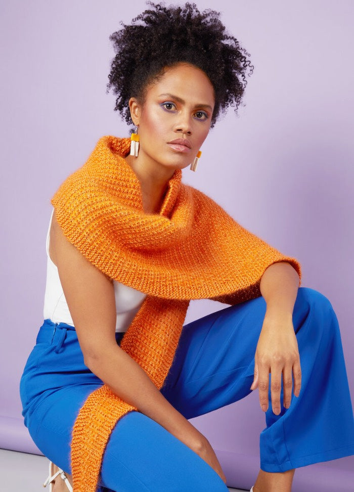 A woman with curly hair poses confidently in front of a lavender background. She wears a textured orange scarf, white top, and vibrant blue pants.