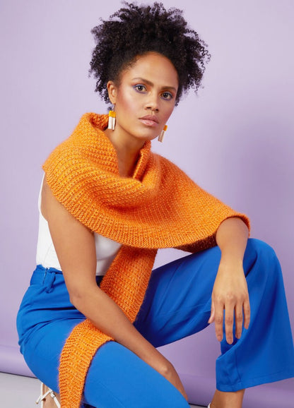 A woman with curly hair poses confidently in front of a lavender background. She wears a textured orange scarf, white top, and vibrant blue pants.