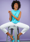 A joyful woman in a bright blue top and white pants sits on a stool against a lavender background. Her vibrant smile conveys happiness and energy.