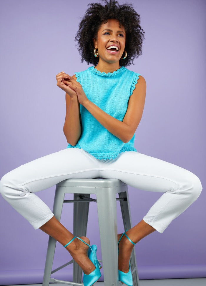 A joyful woman in a bright blue top and white pants sits on a stool against a lavender background. Her vibrant smile conveys happiness and energy.
