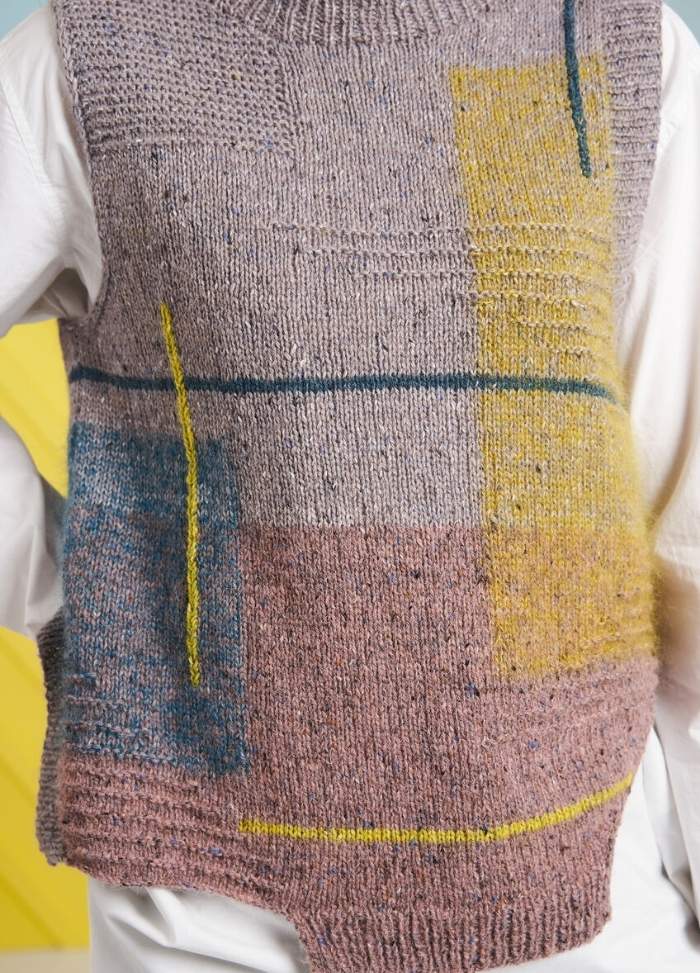 Close-up of a knitted vest featuring abstract patterns in muted pink, blue, and yellow tones, over a white shirt. The background is pastel yellow.