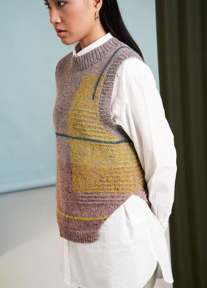 A person wearing a white shirt and a textured, colorful knit vest with geometric patterns. The background is soft blue and green, creating a calm tone.