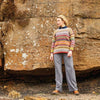 A person stands against a rocky, textured wall, wearing a colorful, patterned sweater and gray pants. The scene conveys a calm, rustic outdoor vibe.
