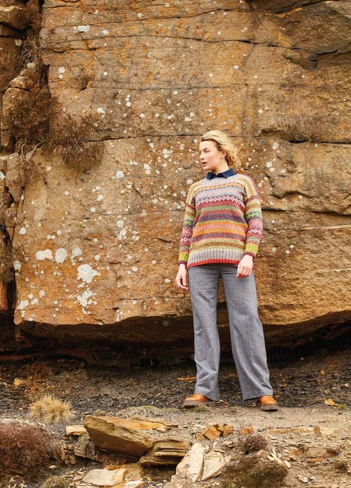 A person stands against a rocky, textured wall, wearing a colorful, patterned sweater and gray pants. The scene conveys a calm, rustic outdoor vibe.