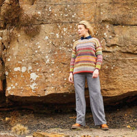 A person stands against a rocky, textured wall, wearing a colorful, patterned sweater and gray pants. The scene conveys a calm, rustic outdoor vibe.
