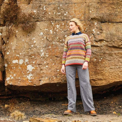 A person stands against a rocky, textured wall, wearing a colorful, patterned sweater and gray pants. The scene conveys a calm, rustic outdoor vibe.