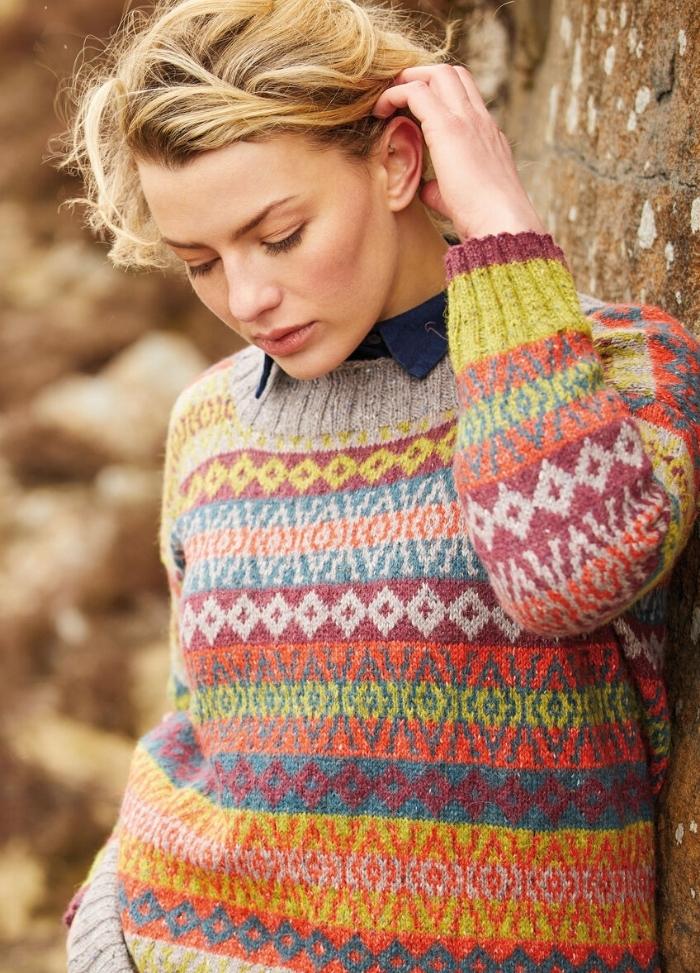 A person with blonde hair leans against a stone wall, wearing a colorful, patterned knit sweater. The expression is thoughtful, creating a calm and introspective mood.