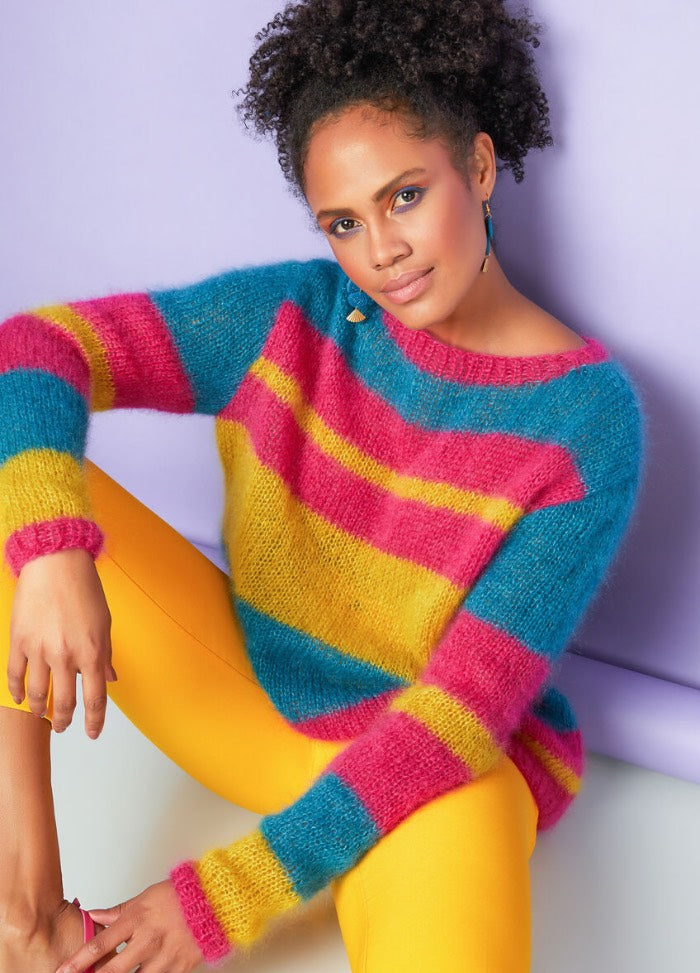 A person sits against a lavender background, wearing a colorful fuzzy sweater with blue, pink, and yellow stripes, paired with bright yellow pants. They have curly hair and a relaxed expression.