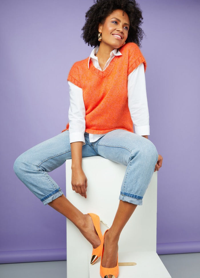 Woman in an orange sweater vest over a white shirt, with rolled-up jeans and orange heels, sits on a white cube against a purple background, smiling confidently.