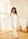 Woman in a flowing white dress and shawl, smiling softly. She stands barefoot on wooden floor, sunlit with a woven basket in the background.