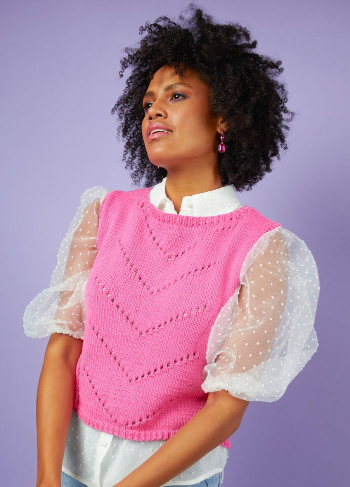 A woman with curly hair wears a bright pink sleeveless knit top over a white blouse with sheer, puffy sleeves, set against a lavender background.