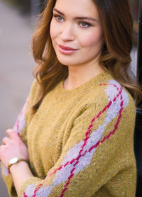 Woman with long brown hair in a mustard sweater with colored patterns, smiling slightly. She stands outdoors with a blurred background, emitting a calm, warm vibe.