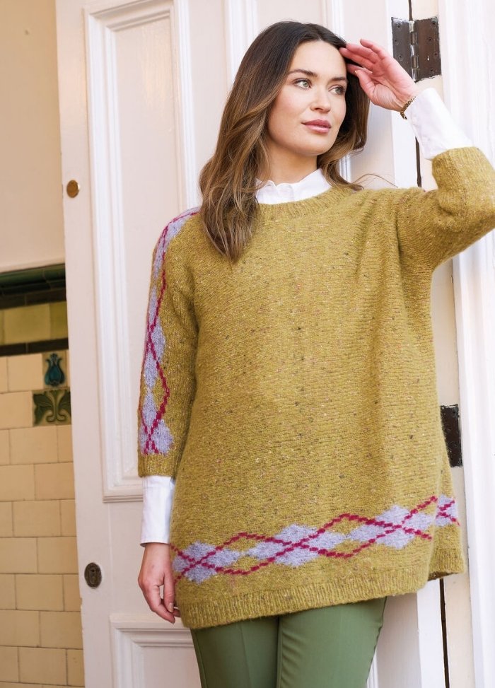 Woman in a cozy mustard sweater with pink and blue patterns stands in a doorway. She looks thoughtful, wearing olive pants and a white shirt.