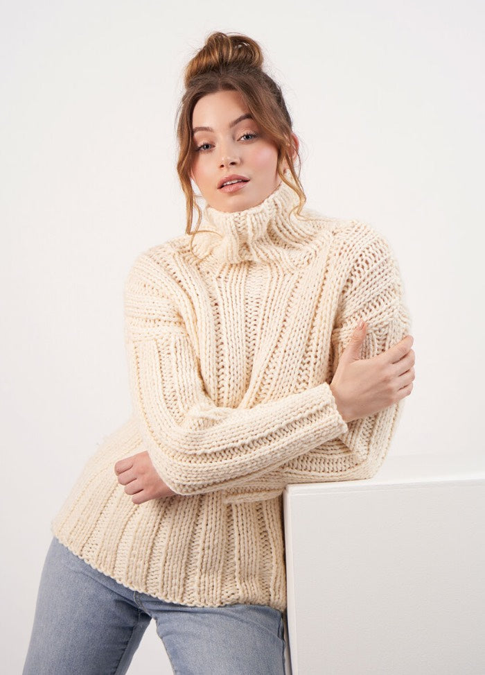 Woman with light brown hair in a bun, wearing a cream chunky knit turtleneck sweater and jeans. She leans casually against a white surface, exuding coziness.
