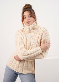 Woman with light brown hair in a bun, wearing a cream chunky knit turtleneck sweater and jeans. She leans casually against a white surface, exuding coziness.