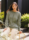 Woman with long dark hair wearing a green sweater and beige pants sits on a yellow chair outdoors. Greenery and a dark building are in the background.