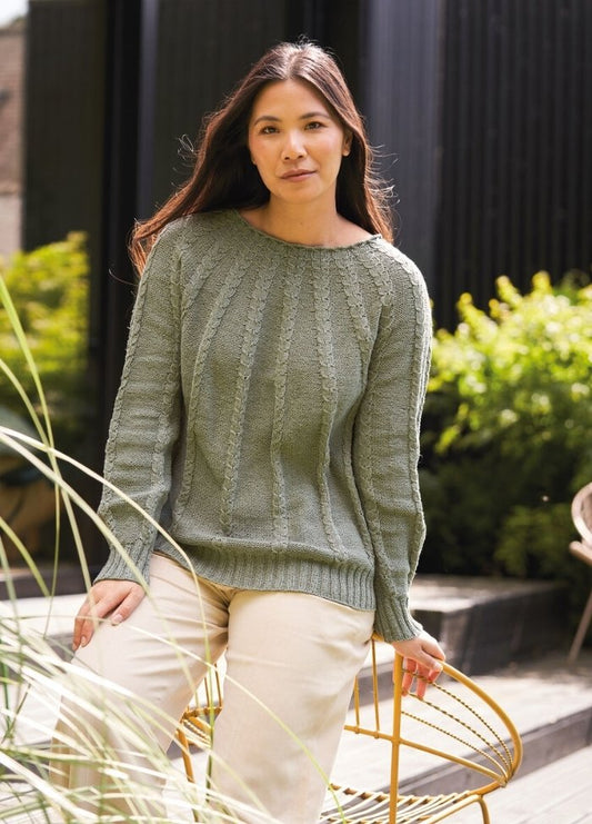 Woman with long dark hair wearing a green sweater and beige pants sits on a yellow chair outdoors. Greenery and a dark building are in the background.