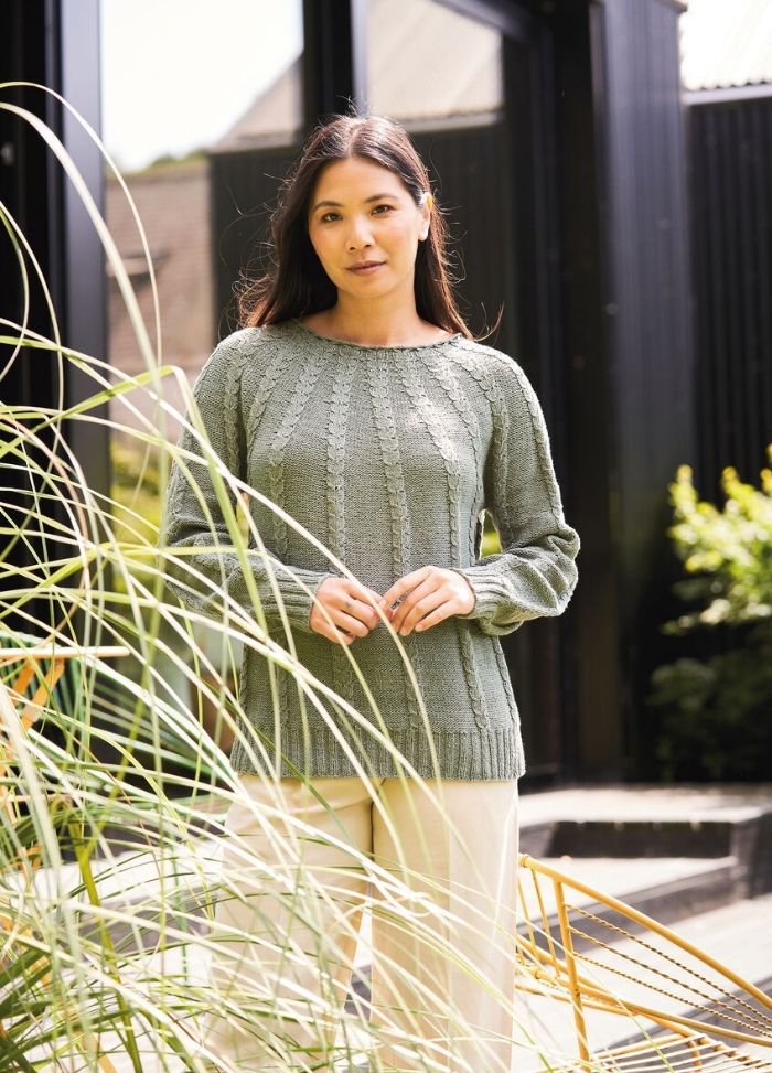 Woman in a green knitted sweater stands outdoors near tall grass, with a modern building in the background. The scene conveys a serene, casual vibe.