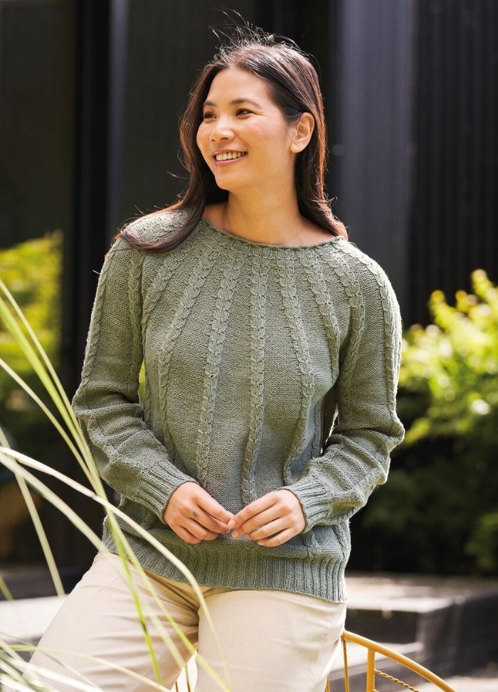 Smiling woman in a green sweater with a textured pattern stands outdoors. She is framed by greenery, conveying a relaxed, cheerful mood.