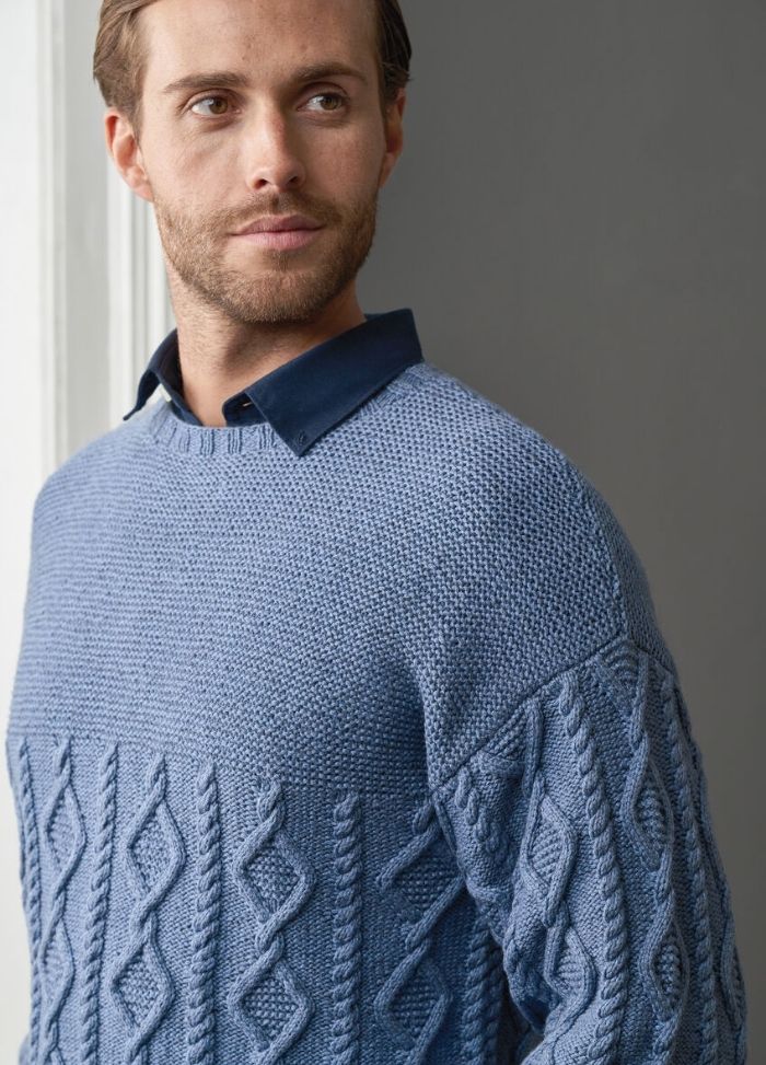 A man with a neatly trimmed beard gazes to the side, wearing a textured blue sweater with intricate patterns over a dark collared shirt, exuding a calm and thoughtful demeanor.