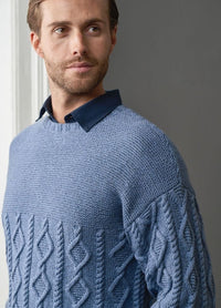 A man with a neatly trimmed beard gazes to the side, wearing a textured blue sweater with intricate patterns over a dark collared shirt, exuding a calm and thoughtful demeanor.