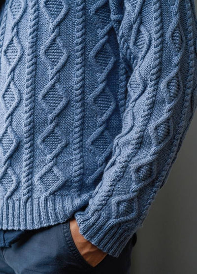 A person wearing a blue cable-knit sweater with intricate patterns and texture, standing with hands in pockets. The tone is cozy and stylish.