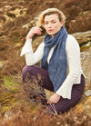Woman in a white sweater and blue scarf sits on rocky terrain, surrounded by brown shrubs. She gazes thoughtfully, evoking a serene, contemplative mood.