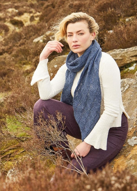 Woman in a white sweater and blue scarf sits on rocky terrain, surrounded by brown shrubs. She gazes thoughtfully, evoking a serene, contemplative mood.