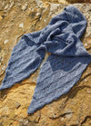 Blue knitted scarf with a diamond pattern, artfully arranged on a sunlit, textured stone surface. The warm light contrasts with the scarf's cool tone.