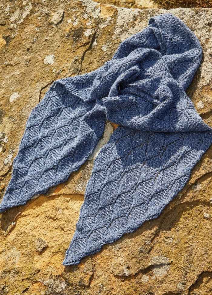 Blue knitted scarf with a diamond pattern, artfully arranged on a sunlit, textured stone surface. The warm light contrasts with the scarf's cool tone.