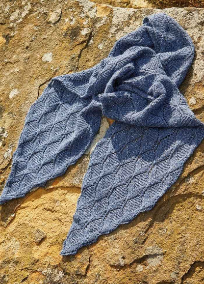 Blue knitted scarf with a diamond pattern, artfully arranged on a sunlit, textured stone surface. The warm light contrasts with the scarf's cool tone.