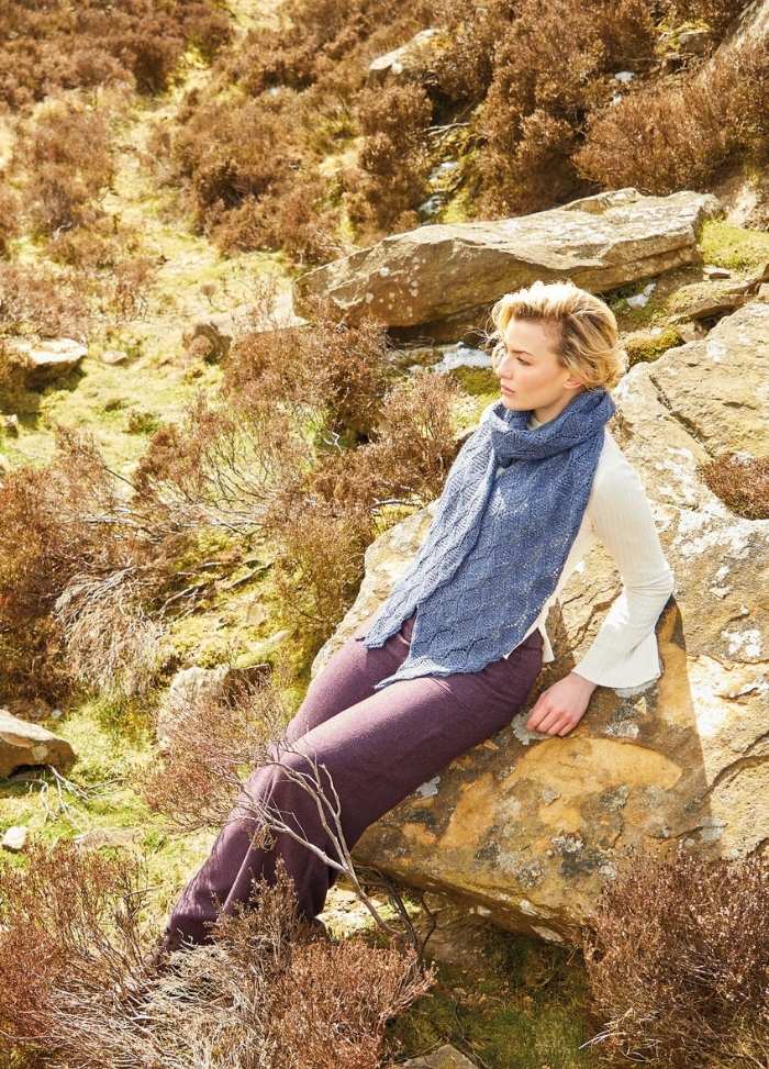 A person with short hair, dressed in a long blue scarf and cream top, relaxes on a rocky hillside. The scene conveys a calm, serene feeling.