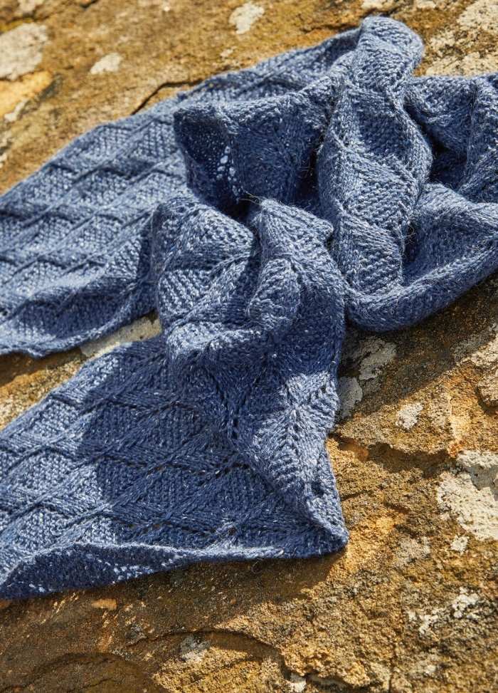 Blue knitted scarf with a diamond pattern, artfully draped on a textured, sunlit rock surface. The scene conveys warmth and coziness.