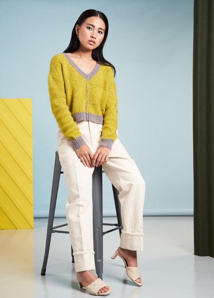 A woman sits on a stool, wearing a yellow and gray sweater, cream pants, and beige heels. The background is light blue with a yellow panel, conveying a calm, stylish tone.