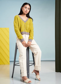 A woman sits on a stool, wearing a yellow and gray sweater, cream pants, and beige heels. The background is light blue with a yellow panel, conveying a calm, stylish tone.