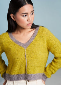 Young woman in a yellow knit sweater with gray trim stands against a light blue background, looking thoughtfully to the right, conveying a calm demeanor.
