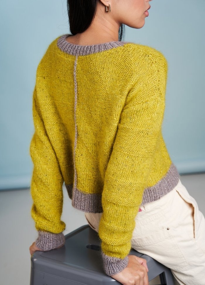 A woman in a yellow sweater with gray accents leans against a gray box, facing away. The background is a soft blue tone, creating a calm atmosphere.