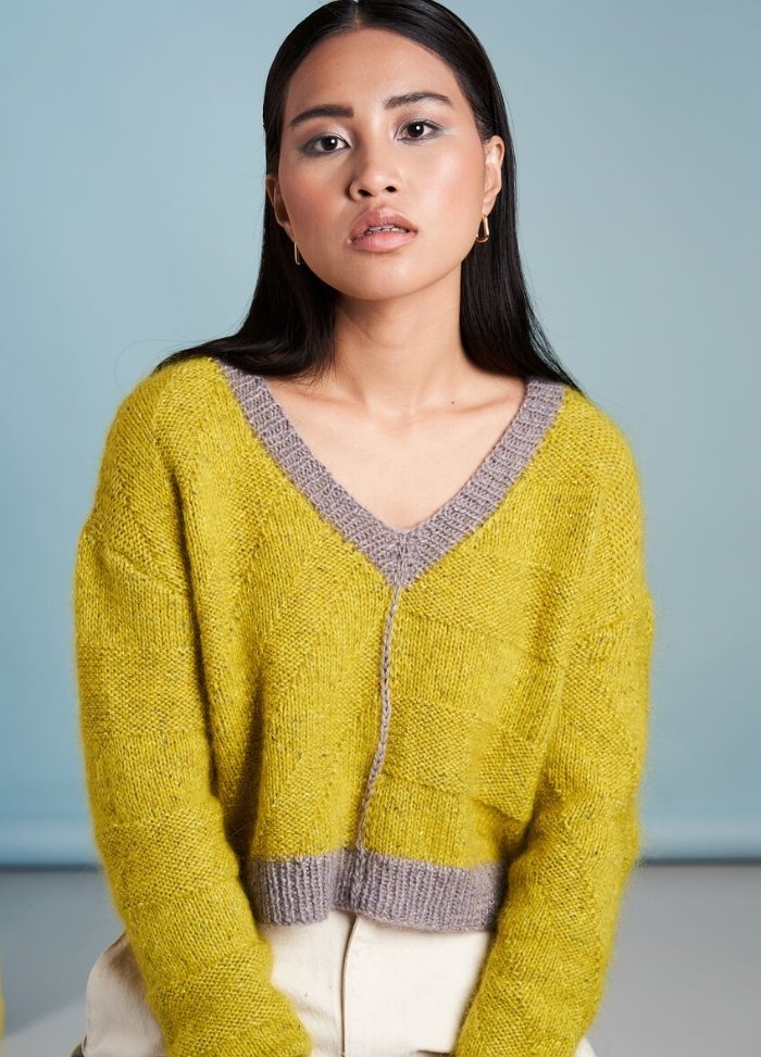 A person with long dark hair wearing a yellow sweater with gray trim sits against a light blue background, displaying a calm, contemplative expression.