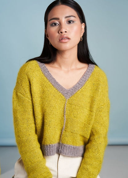A person with long dark hair wearing a yellow sweater with gray trim sits against a light blue background, displaying a calm, contemplative expression.