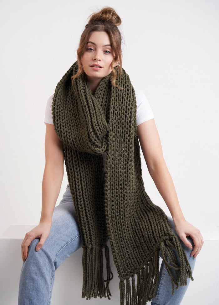 Woman sitting on a white bench, wearing a long, chunky olive-green knit scarf over a white T-shirt and light blue jeans. Relaxed and casual vibe.